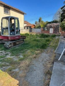 Mini pelleteuse Takeuchi TB153FR stationnée dans un jardin en chantier avec une échelle métallique à droite.