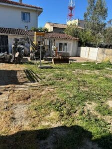 Mini-pelle Volvo dans un jardin en travaux devant une maison blanche avec pylône de télécommunication au loin.